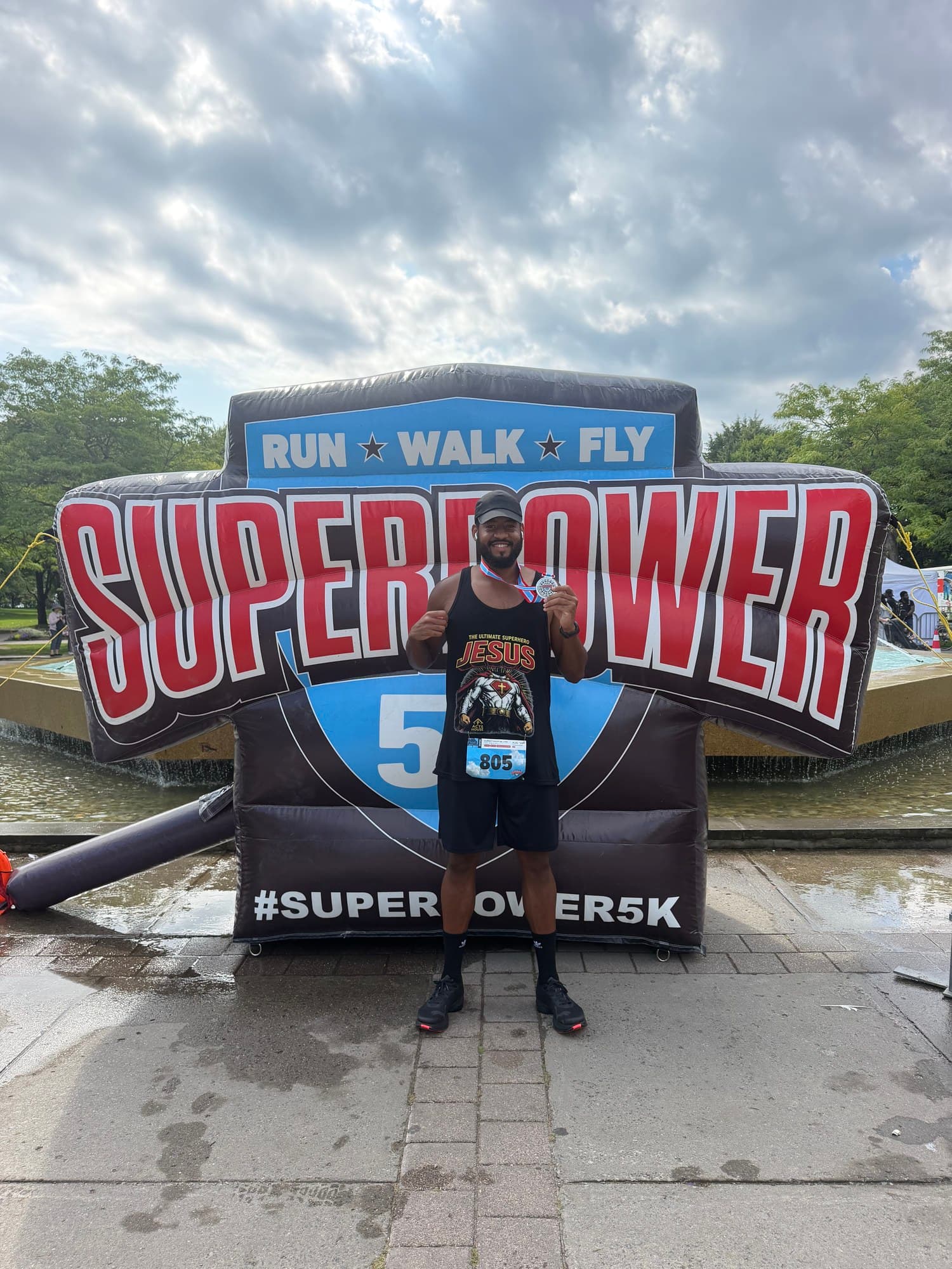Tiye Martin holding up a finisher’s medal after a 5K, wearing a shirt that reads “The Ultimate Superhero Jesus”.